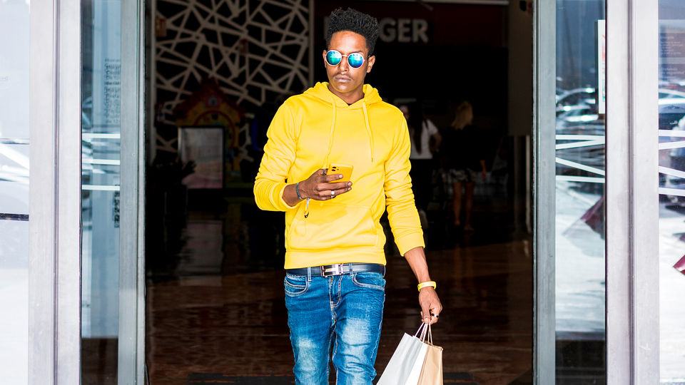 stylish young man in sunglasses yellow sweater and jeans leaving retail store carrying shopping bag holding cellphone