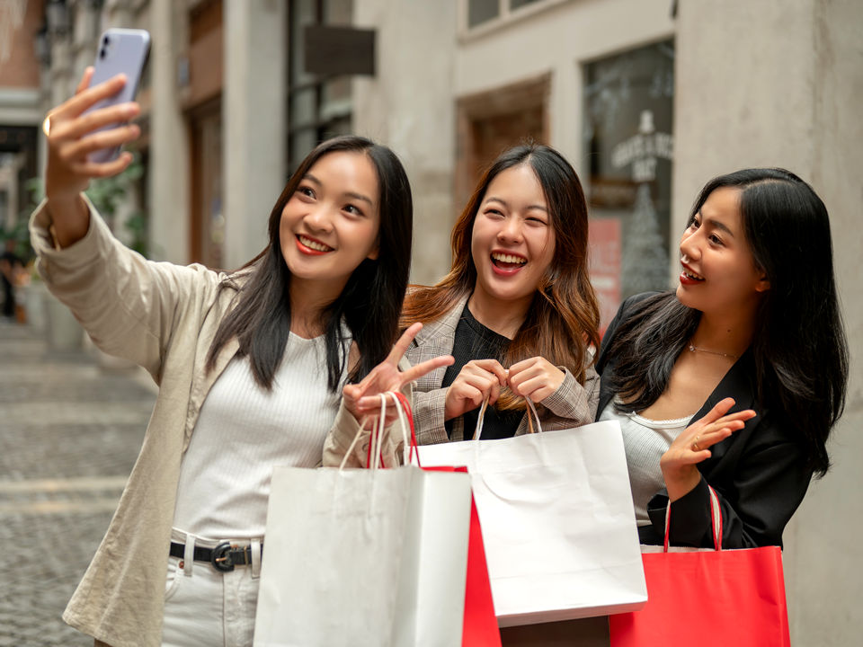 three young female shoppers carrying shopping bags outside retail store taking selfie on smartphone