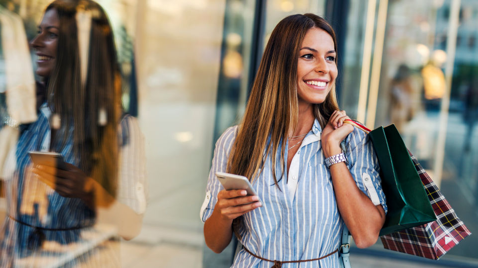 female shopper carrying several shopping bags and her smartphone looking in a retail store window