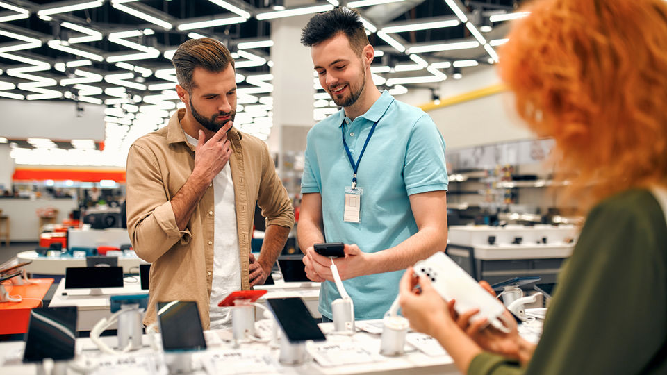 male retail sales associate assistant helps male client shopping for mobile phones while female customer browses 