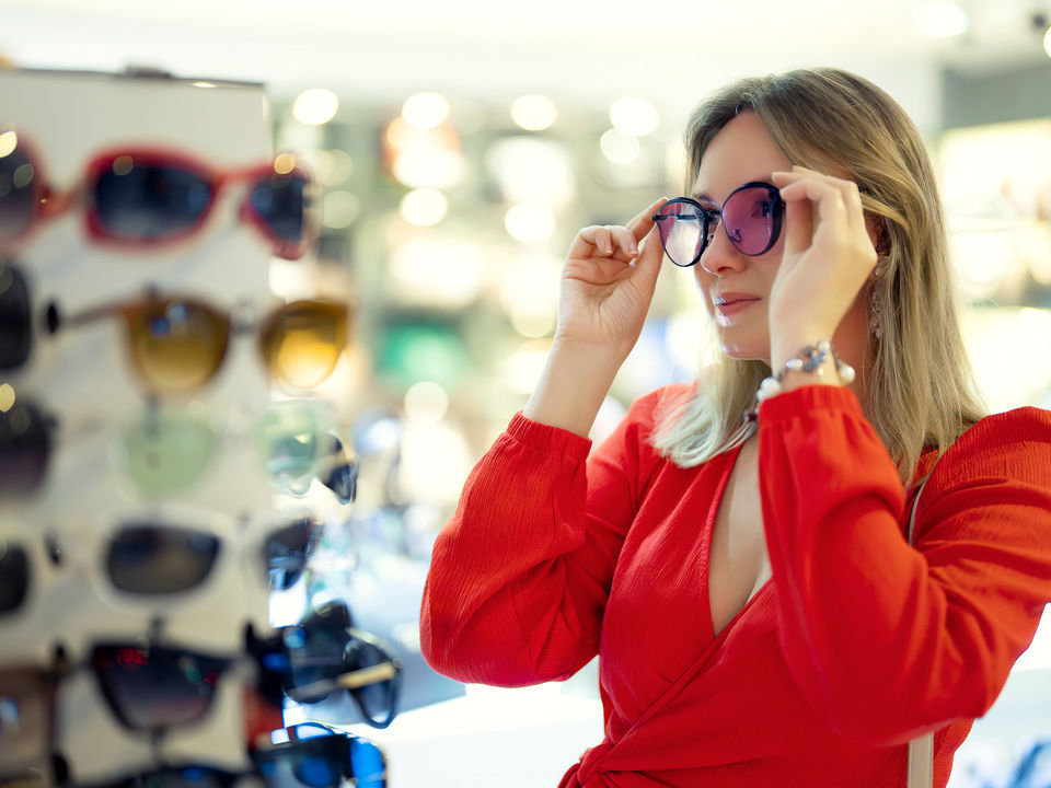 female shopper in brght red dress looking in mirror and trying on a pair of sunglasses in a retail optical store