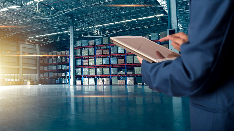 retail professional standing in spacious naturally-lit warehouse reviwing inventory on electronic device tablet