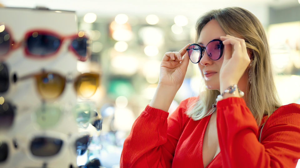 female shopper in brght red dress looking in mirror and trying on a pair of sunglasses in a retail optical store