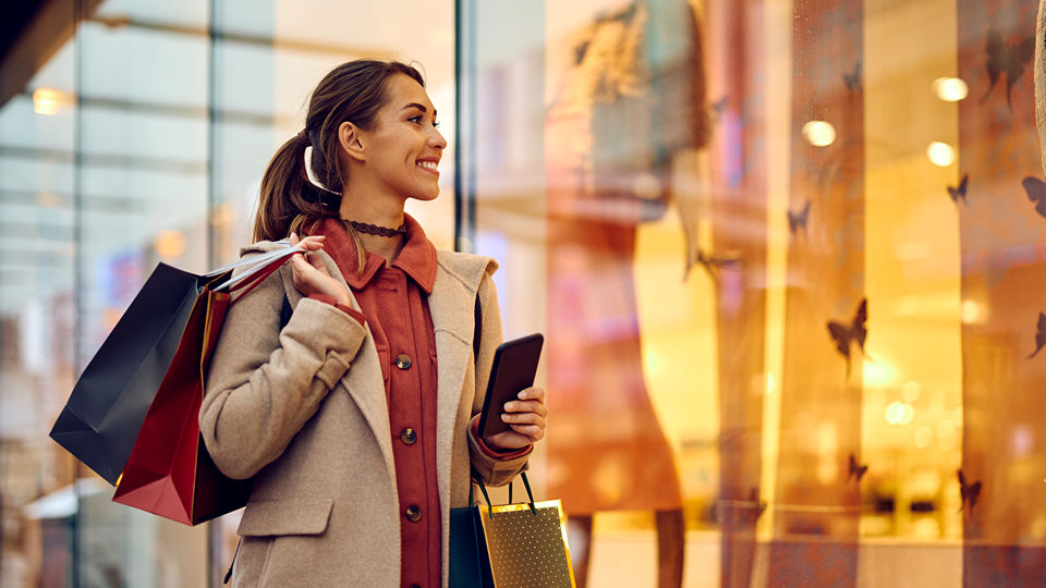 female shopper carrying shopping bag and admiring warmly lit retail store window holiday displays