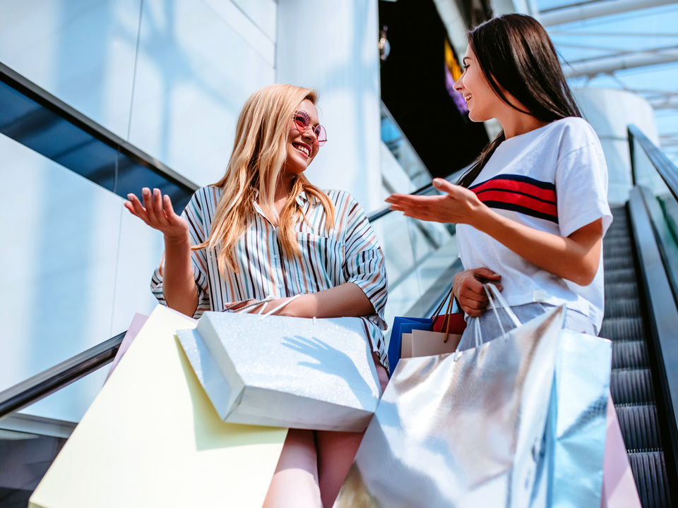 2 female shoppers carrying shopping bags riding an escalator in a retail shopping mall